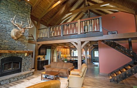 View of living room/kitchen and lofted area with a railing with the vertical slats designed to look like paddles. Large stone fireplace with large Elk mount above.