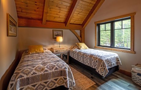 Bedroom with two single beds separated by a night stand. Wood ceiling and large window with black trim.